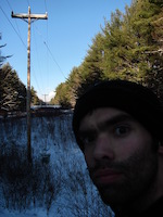 a badly-lit photo of me in front of power lines receding through a cut in the woods in winter